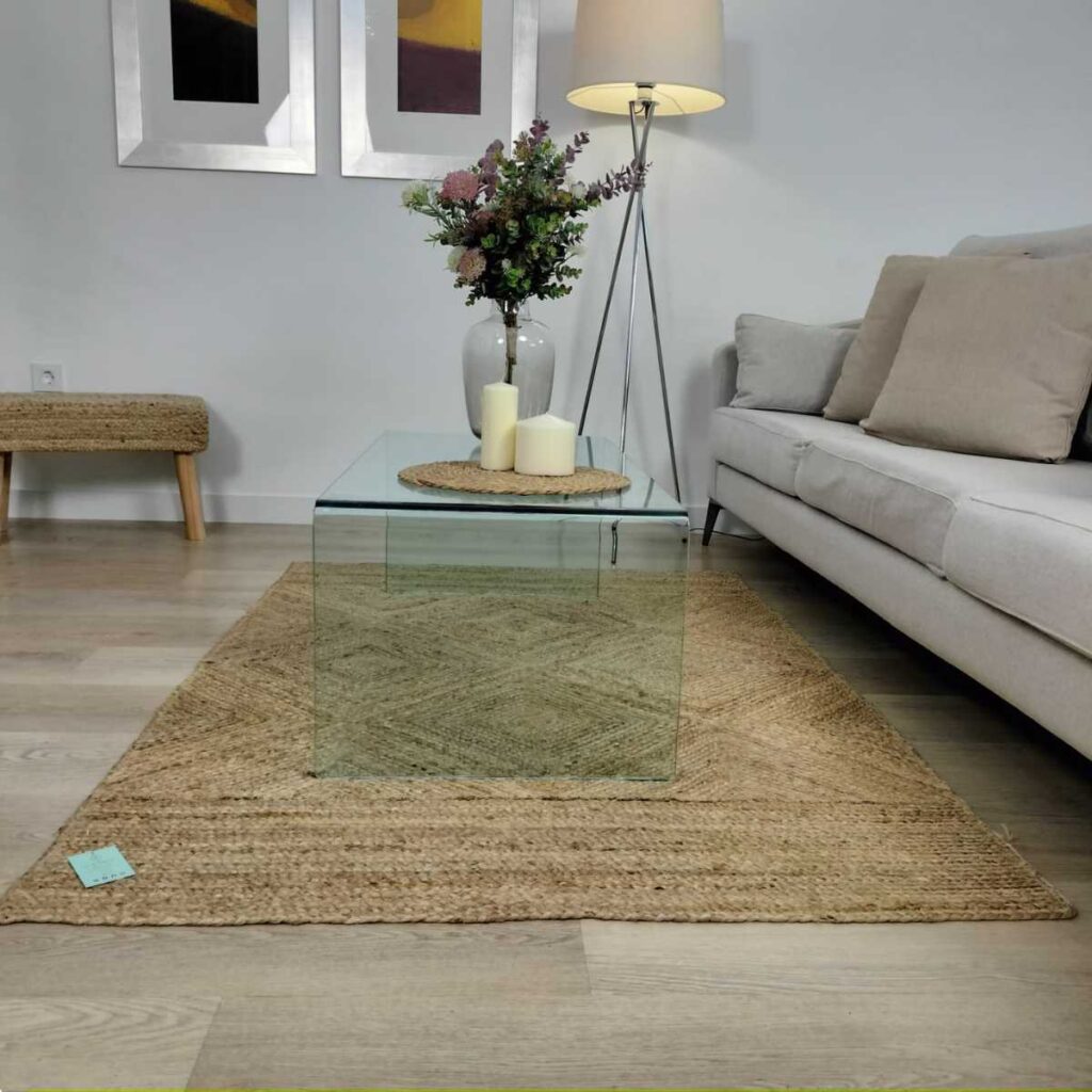 Salón con alfombra de fibras naturales bajo mesa baja de cristal frente a sofá en tonos grises mientras en la mesa hay un pequeño salvamanteles de fibras con velas blancas y al fondo un banco de madera y yute del catálogo de oxiiro